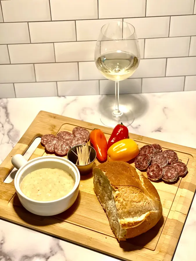 A charcuterie board with sliced sausage, peppers, dip and bread beside a glass of white wine