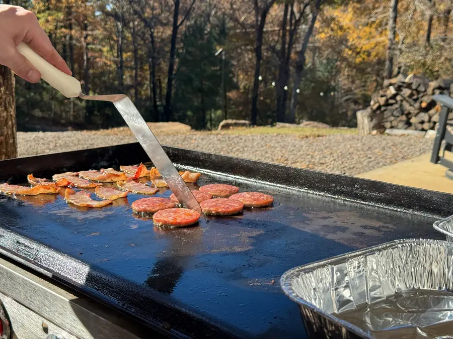 A spatula turning food on an outdoor flat-top griddle, autumn woods behind