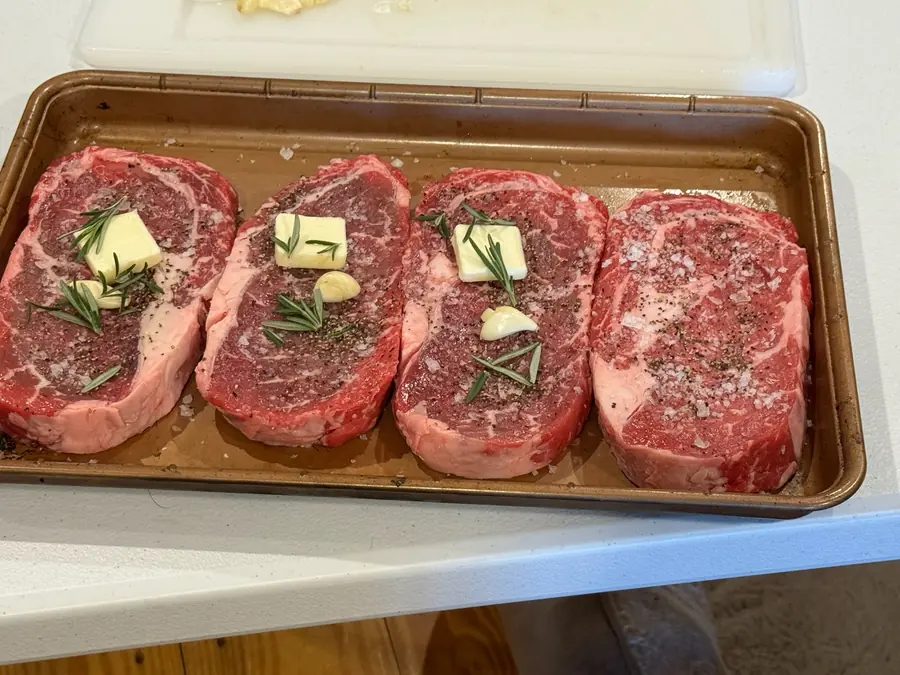 Four seasoned ribeye steaks topped with rosemary and butter on a tray